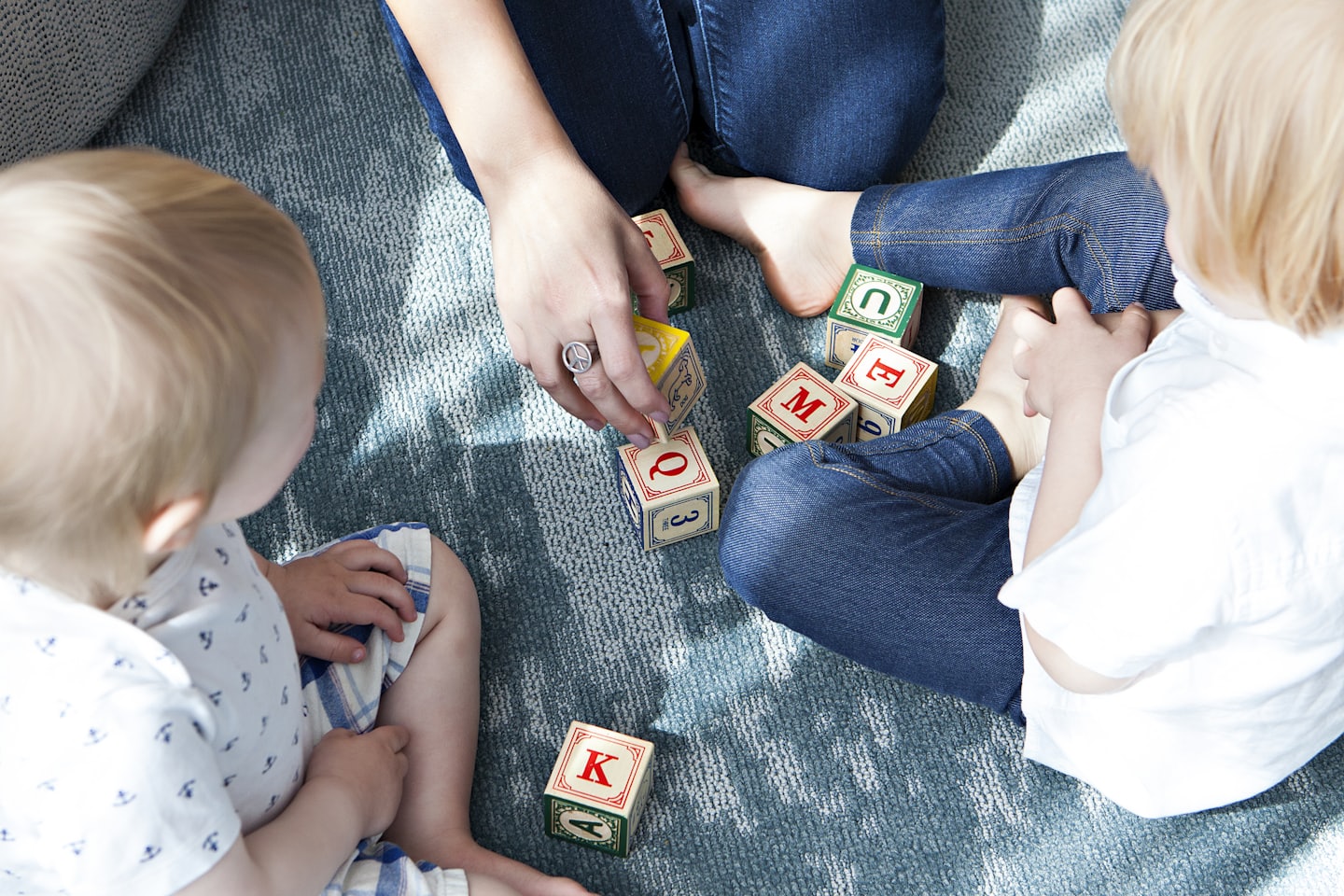 Symbolbild für Bildung und gemeinsames Lernen: Kinder sitzen mit Bezugsperson auf einem Teppich und üben Gemeinsam mit Buchstabenklötzen.