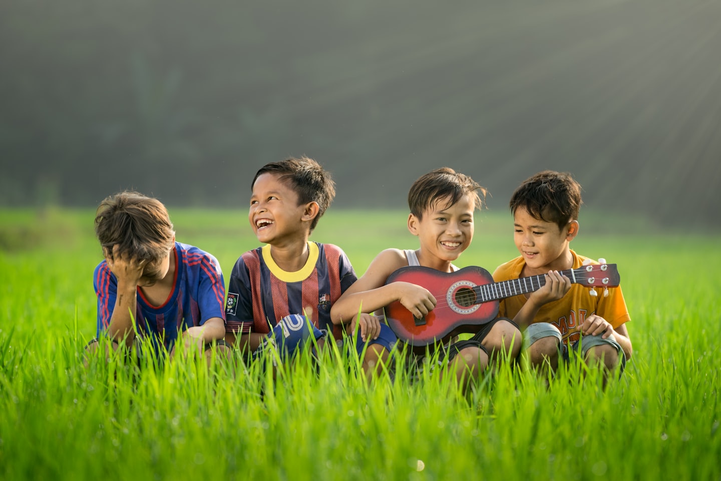 Symbolbild für Freude - vier Kinder sitzen im Grass, genießen die Sonne und lachen gemeinsam. 